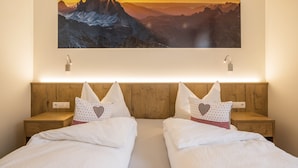 Habitación de hotel con cabecera de madera, dos camas con ropa blanca, almohadas con estampado de corazones y un cuadro grande de un paisaje montañoso sobre las camas.
