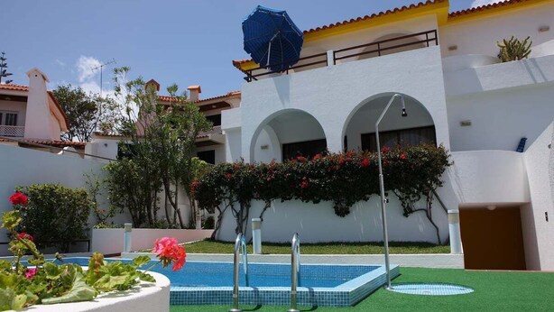 Uma área de piscina com grama sintética, um guarda-sol azul e um edifício branco com arcos.