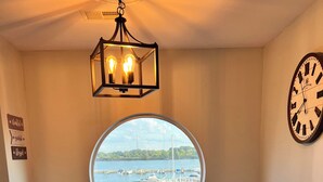 A dining area with a round table, two white chairs, a chandelier, a clock, and a view of a marina through a large window. - Waterfront Marina View Condo - Walk to Jet Express & Downtown Port Clinton (Port Clinton)
