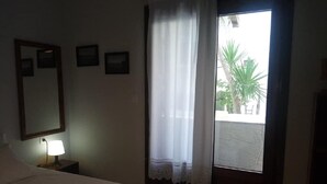 A bedroom with a bed, a nightstand with a lamp, a window with a view of palm trees, and a door leading outside. - Petrino1880 Sea-vacation Apartment in Chalkidiki (Sithonia)