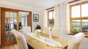 A dining area with a wooden table, white chairs, and a floral table runner. There is a window with curtains and a view of the outdoors. - Osborne Steading (St. Andrews)