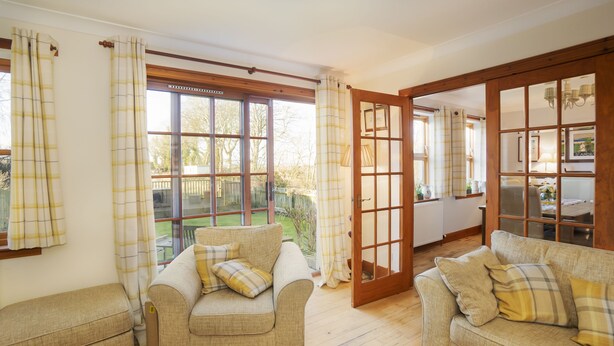 A cozy room with two sofas, a wooden-framed door, and a view of a garden through large windows. - Osborne Steading (St. Andrews)