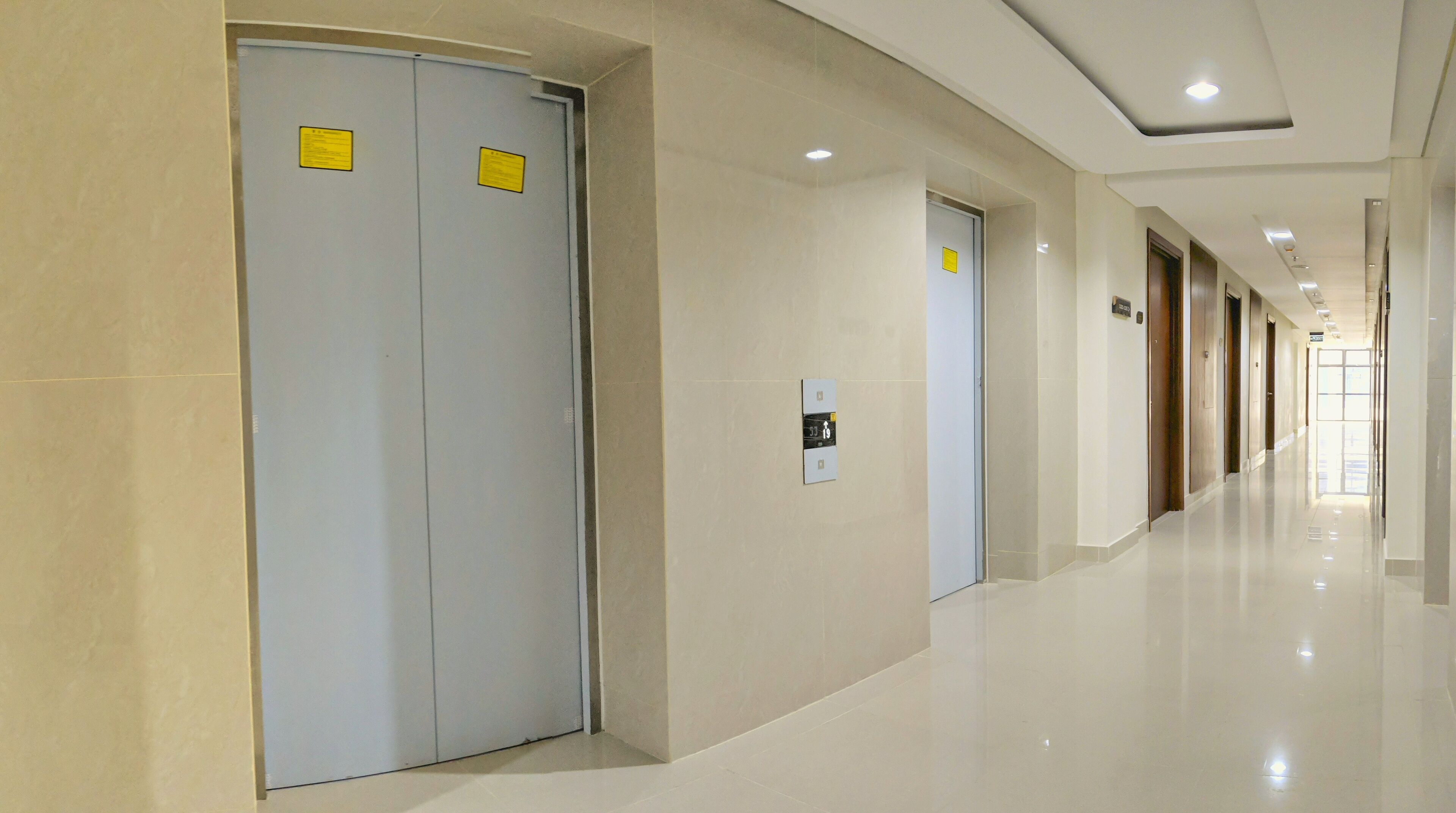 A modern hotel corridor with a shiny floor, double doors with yellow signs, and a clear view of the hallway leading to a bright exit.