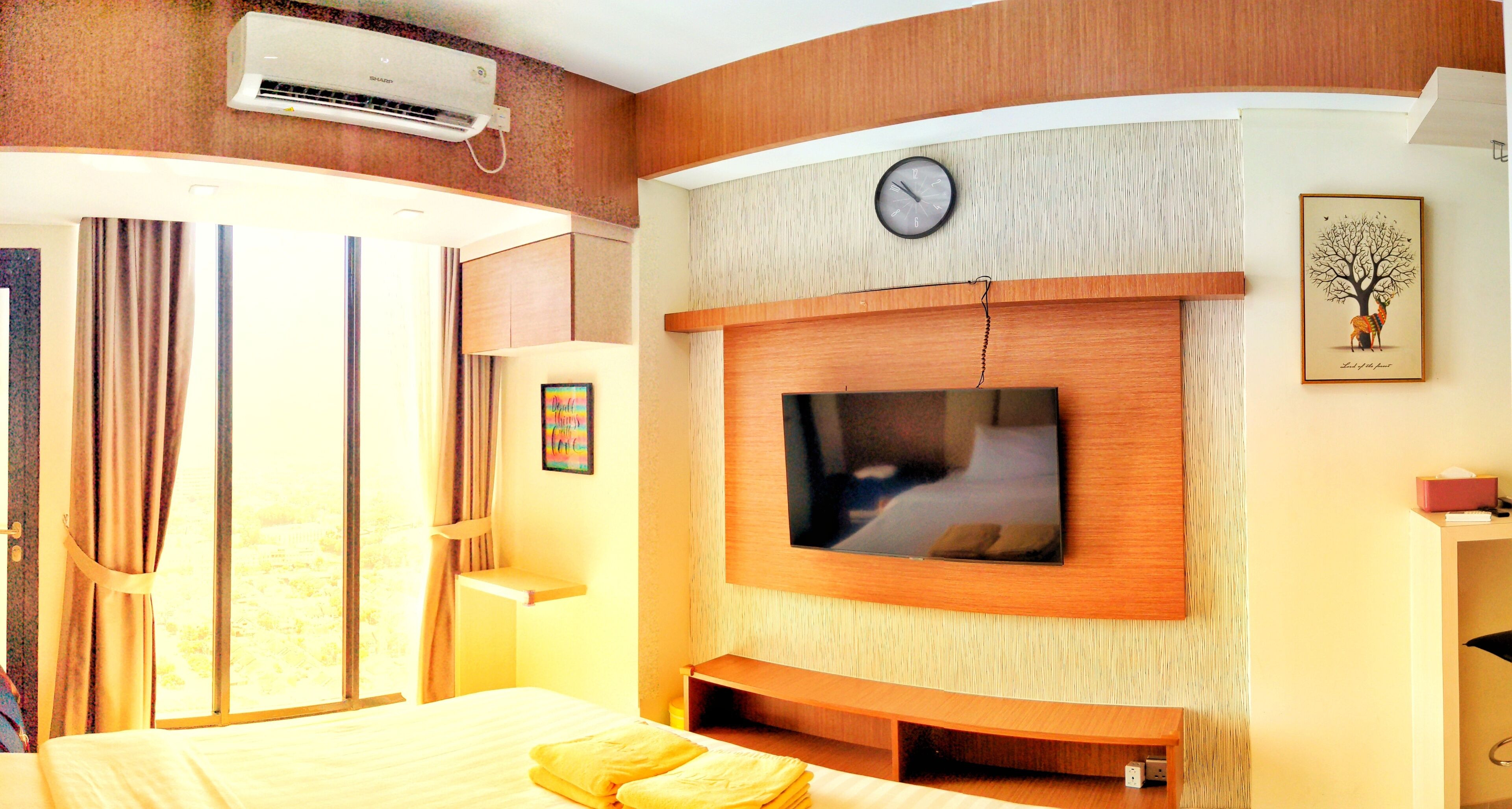 A hotel room with a bed, a TV mounted on the wall, a clock, and a window with curtains.