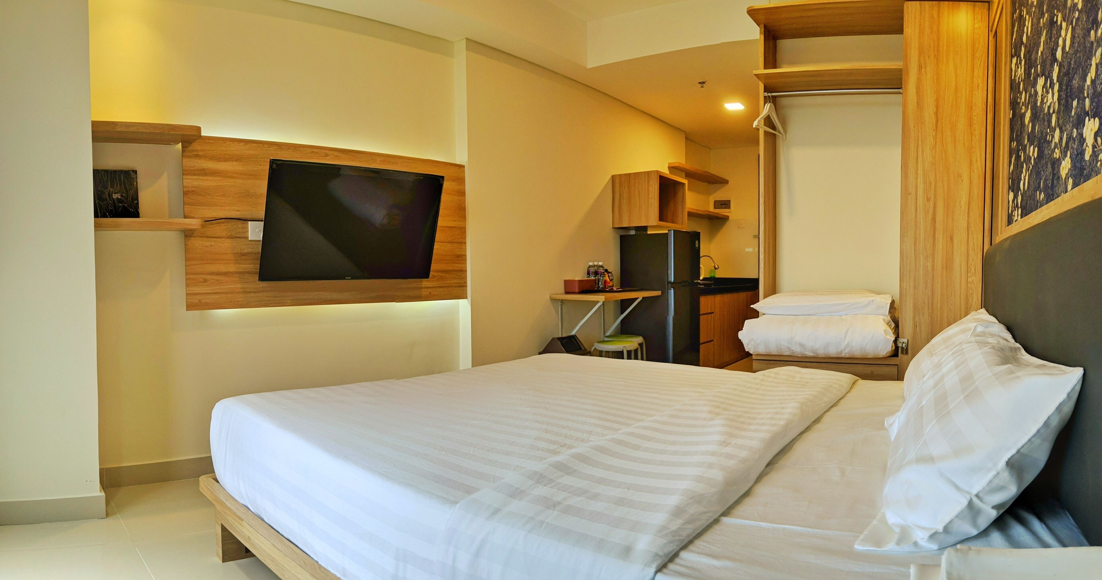 A modern hotel room with a large bed, a flat-screen TV mounted on the wall, a small kitchenette, and a wooden headboard.
