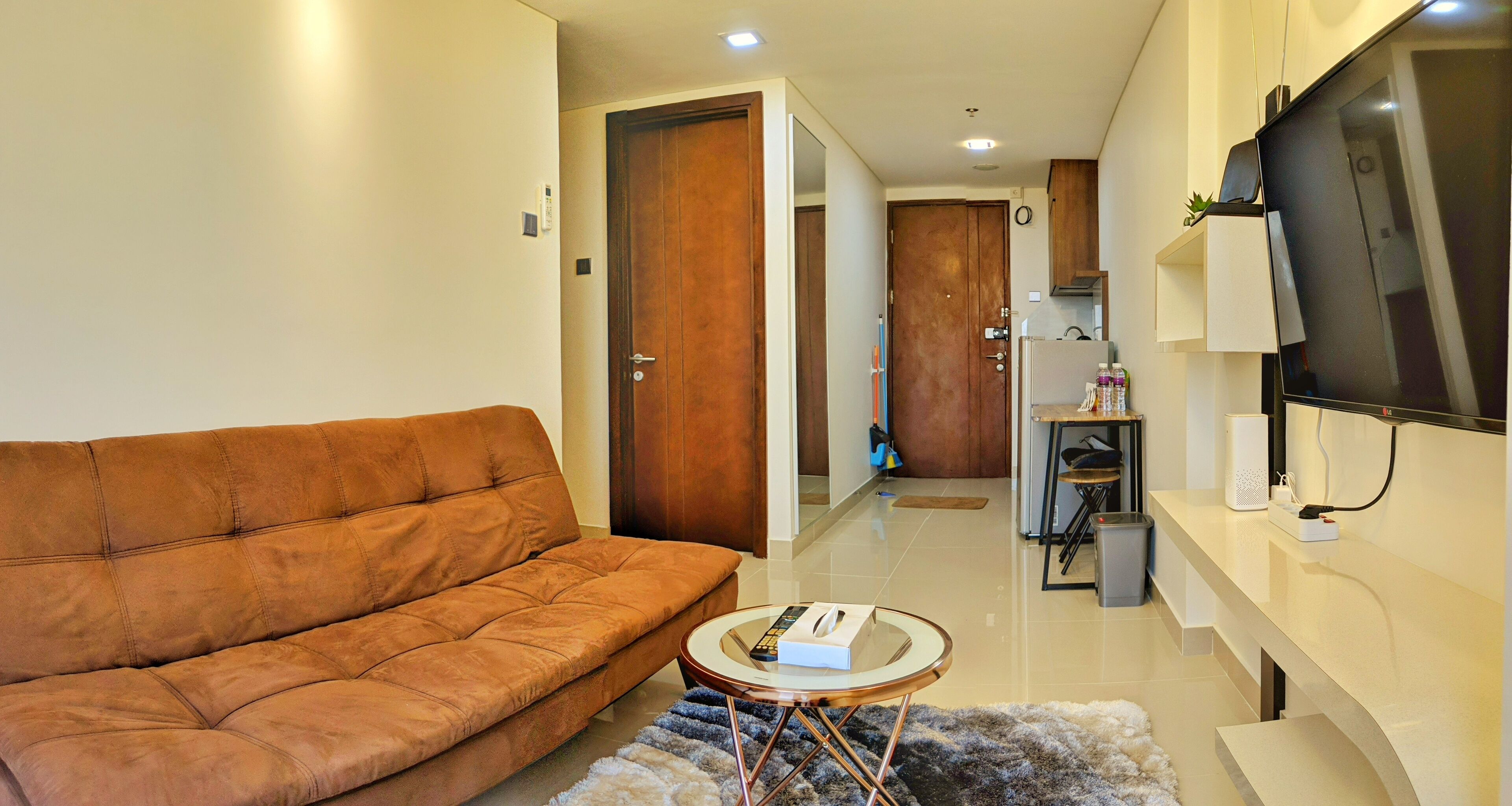 A living room with a brown sofa, a glass coffee table, and a TV mounted on the wall.