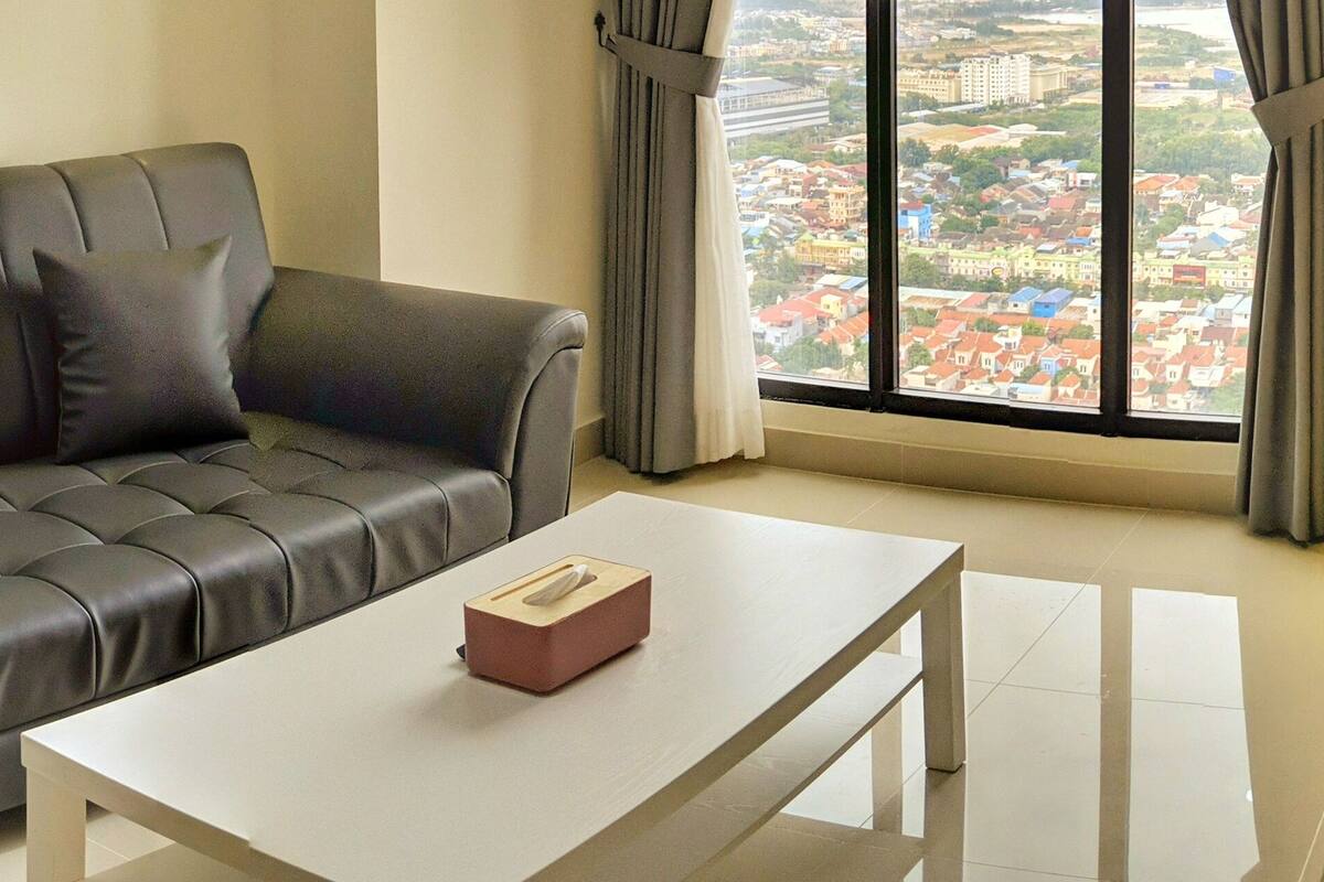 Un salon moderne avec un canapé en cuir, une table basse blanche et une vue sur le paysage urbain grâce à de grandes fenêtres.