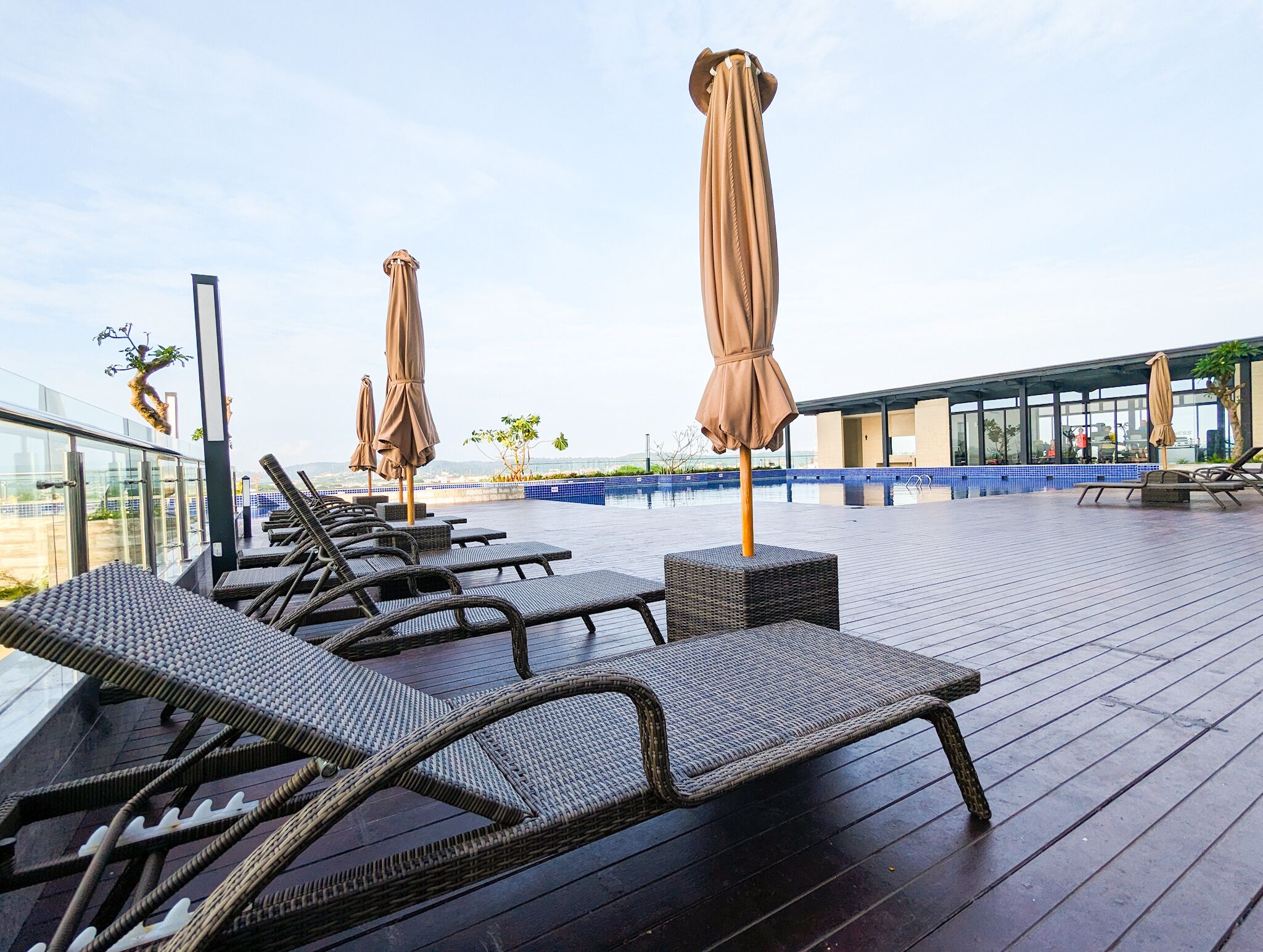A rooftop pool area with lounge chairs and umbrellas, a glass balustrade, and a view of the cityscape.
