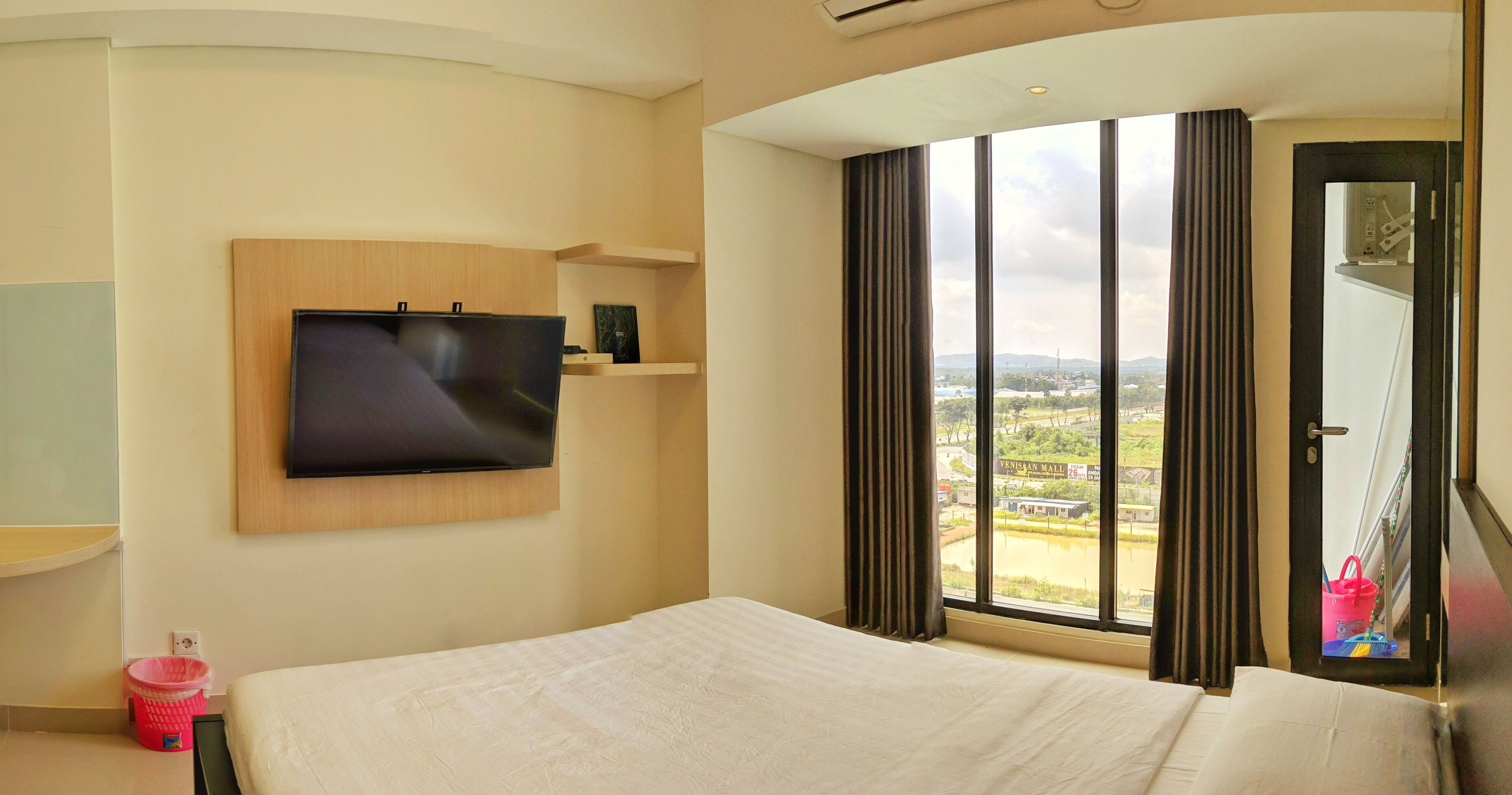 A hotel room with a bed, a TV mounted on the wall, a window with a view of a distant building, and a door with a mirror.
