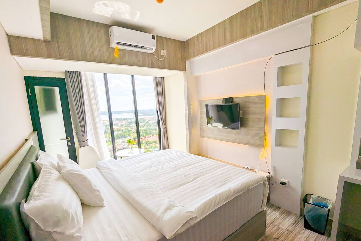 Une chambre d’hôtel moderne dotée d’un grand lit, d’un téléviseur à écran plat fixé au mur et offrant une vue sur la ville grâce à de grandes fenêtres.