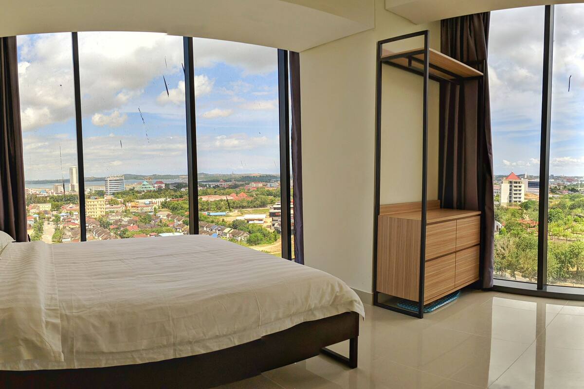 Une chambre à coucher avec un grand lit, une commode en bois et une vue sur le paysage urbain, visible à travers de grandes fenêtres.