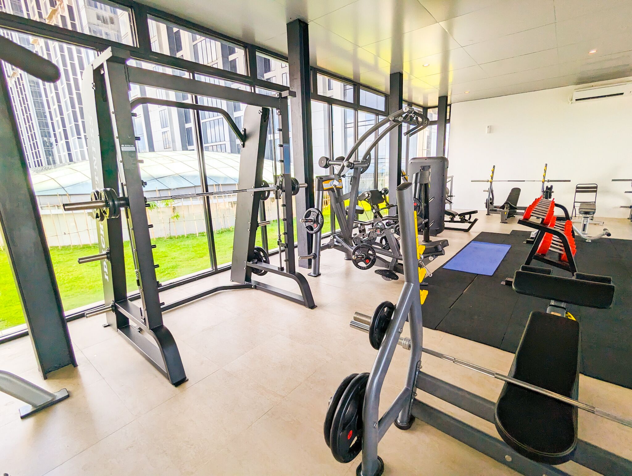 Un gimnasio con diferentes equipos de ejercicio, amplias ventanas y vistas a un área verde exterior.