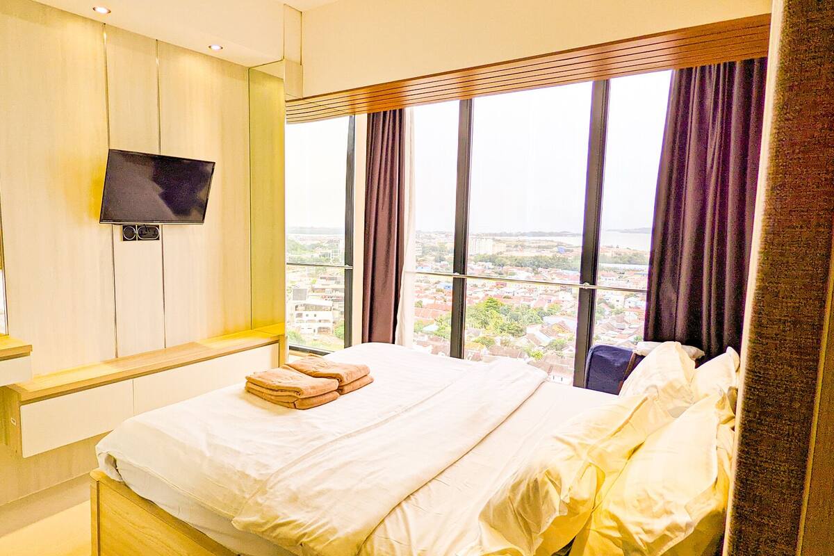 Une chambre d’hôtel avec un lit, un téléviseur fixé au mur et une grande fenêtre donnant sur la ville.