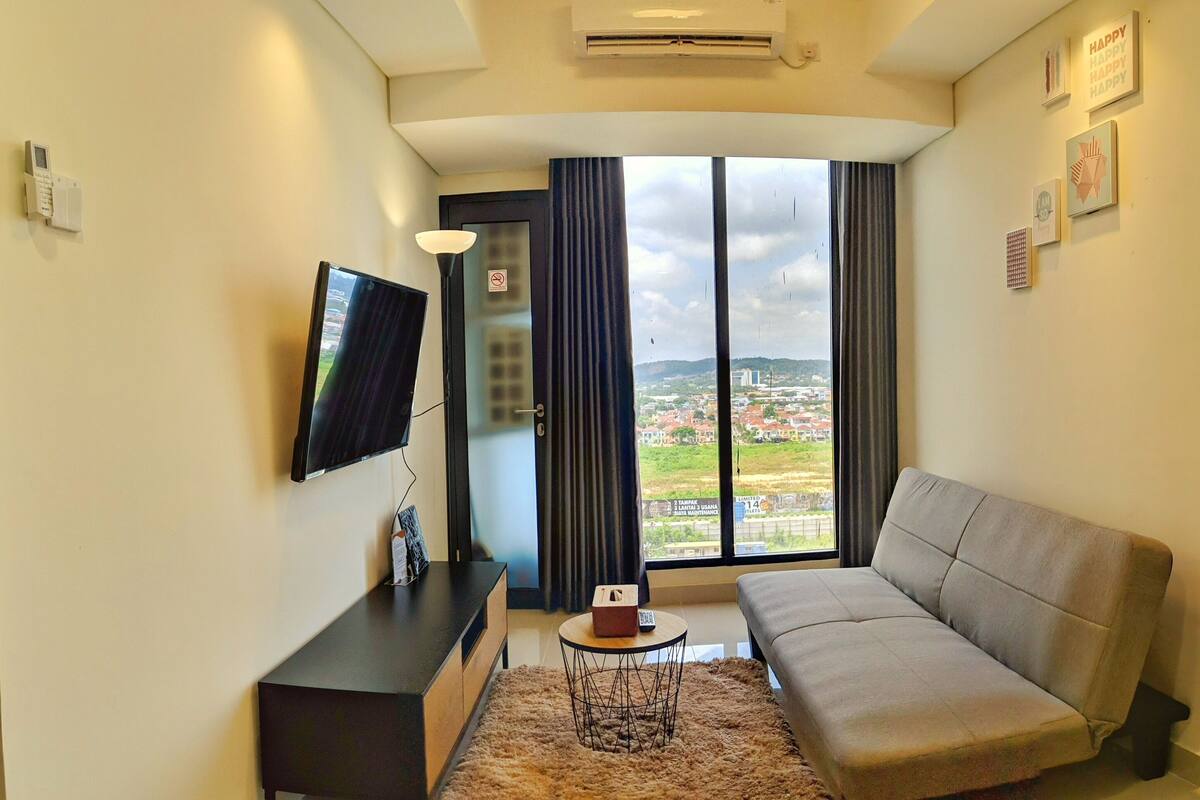 Un salon moderne avec un canapé, un téléviseur, une petite table avec une télécommande et une grande fenêtre donnant sur la ville.