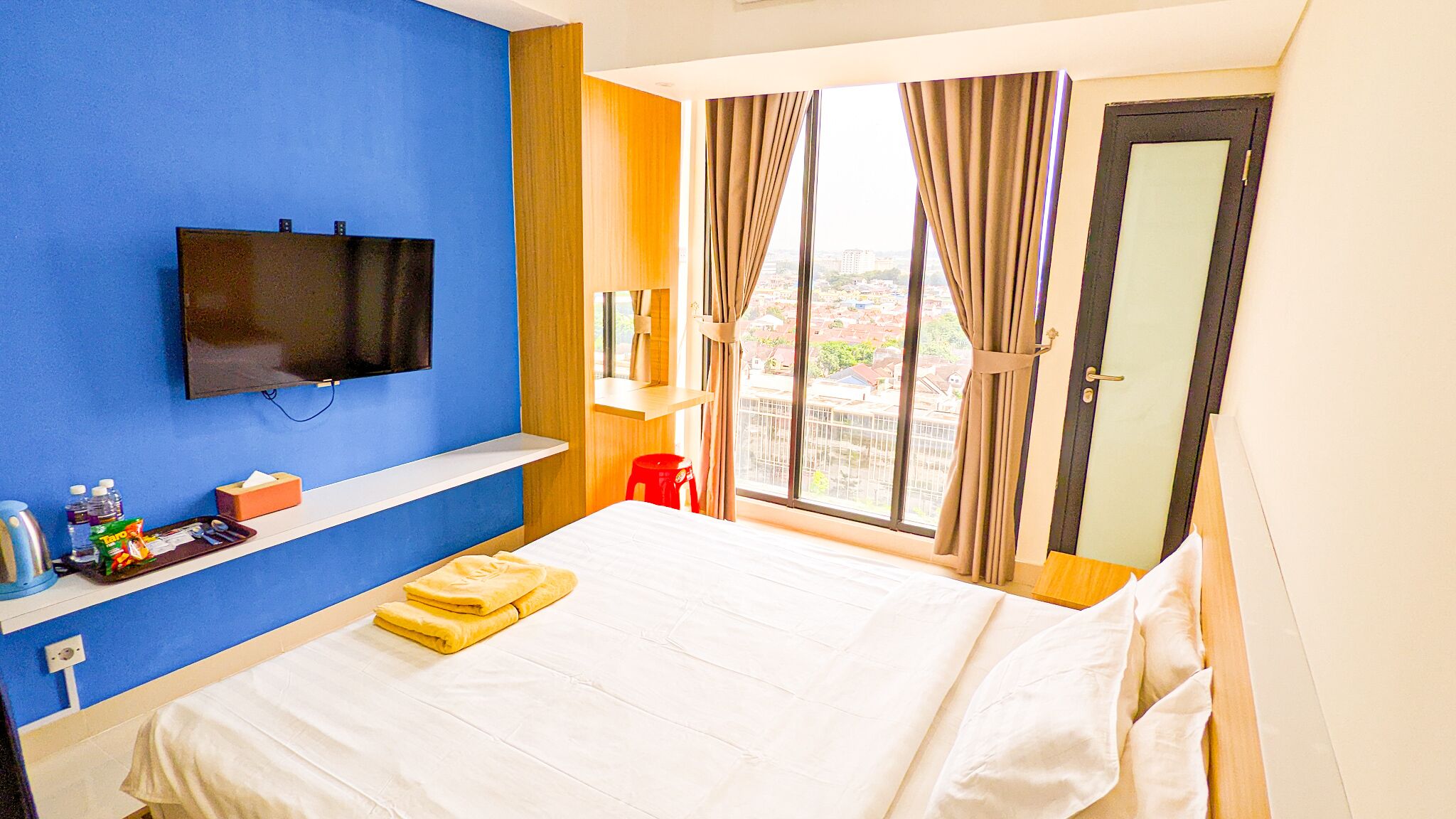 A hotel room with a bed, a TV mounted on the wall, a window with curtains, and a view of the cityscape.