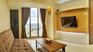A modern hotel room with a large window, a flat-screen TV mounted on a wooden panel, a brown sofa, and a small wooden table with a remote control. - Lovina at Pollux Meisterstadt (Batam)