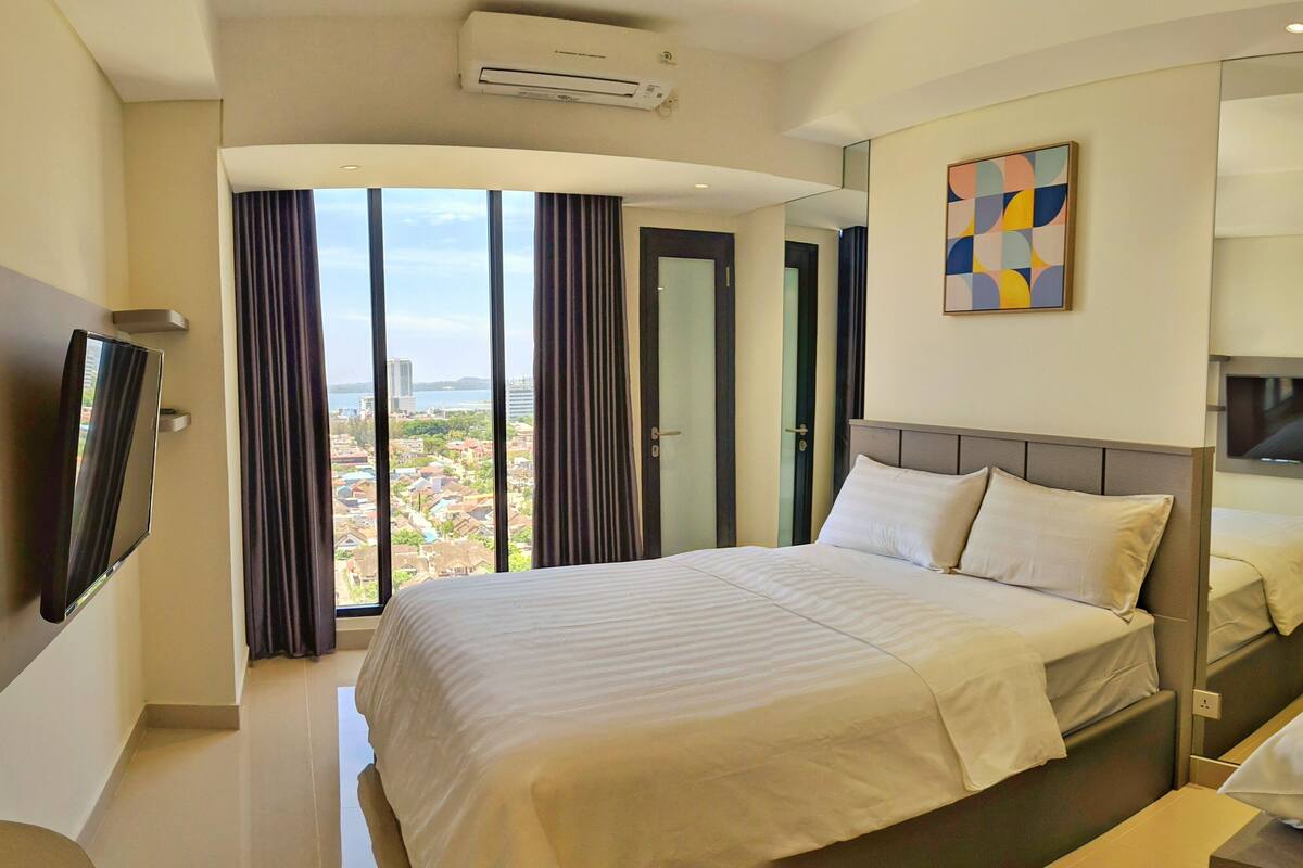 Une chambre d’hôtel dotée d’un grand lit, d’un téléviseur à écran plat fixé au mur, offrant une vue sur la ville depuis la fenêtre, et d’un appareil de climatisation installé au plafond.