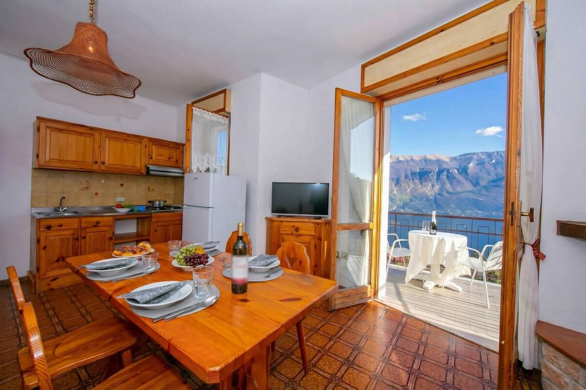 A wooden dining table set with plates, glasses, and a bottle, a kitchen with wooden cabinets, and a balcony with a table and chairs overlooking a mountainous landscape.