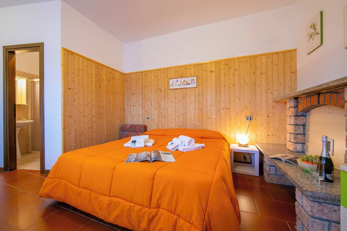 A room with a wooden paneled wall, a bed with an orange bedspread, a small table with a lamp, and a stone fireplace.