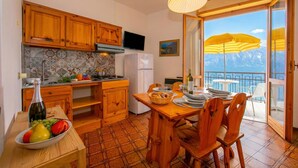 A kitchen with wooden cabinets, a dining table with chairs, a fruit bowl, and a view of mountains through a balcony door. - Appartamenti Secrel (Tremosine)