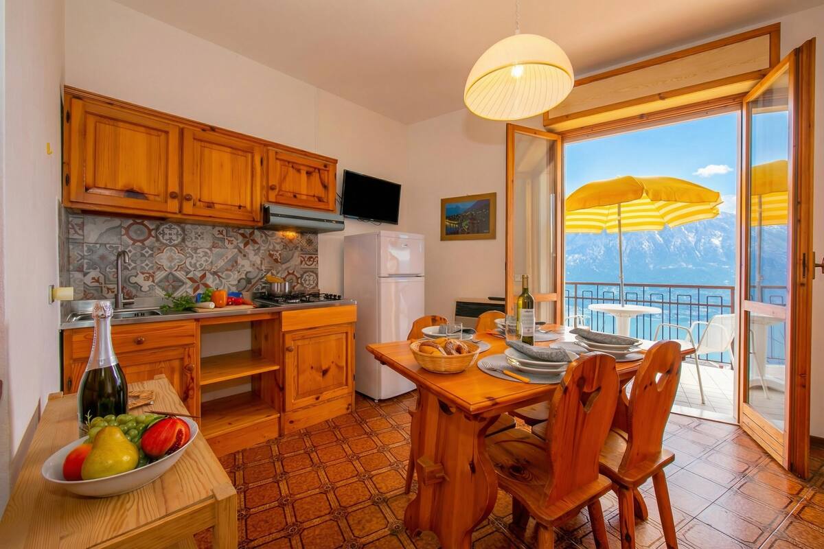A kitchen with wooden cabinets, a dining table with chairs, a fruit bowl, and a view of mountains through a balcony door.