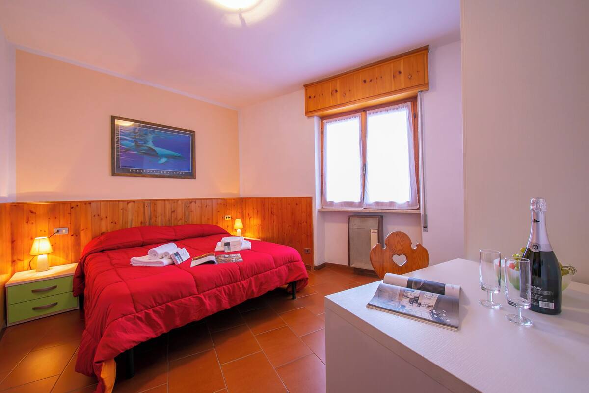 A hotel room with a wooden headboard, a bed with red bedding, a nightstand with a lamp, a window with curtains, and a table with a bottle and glasses.