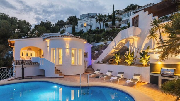 A pool area with a circular pool, lounge chairs, and a staircase leading to a white building with illuminated windows. - Villa INFINIDAD by Abahana Villas (Moraira)