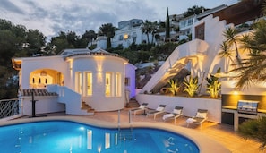 Uma área de piscina com piscina circular, espreguiçadeiras e uma escadaria que leva a um edifício branco com janelas iluminadas.