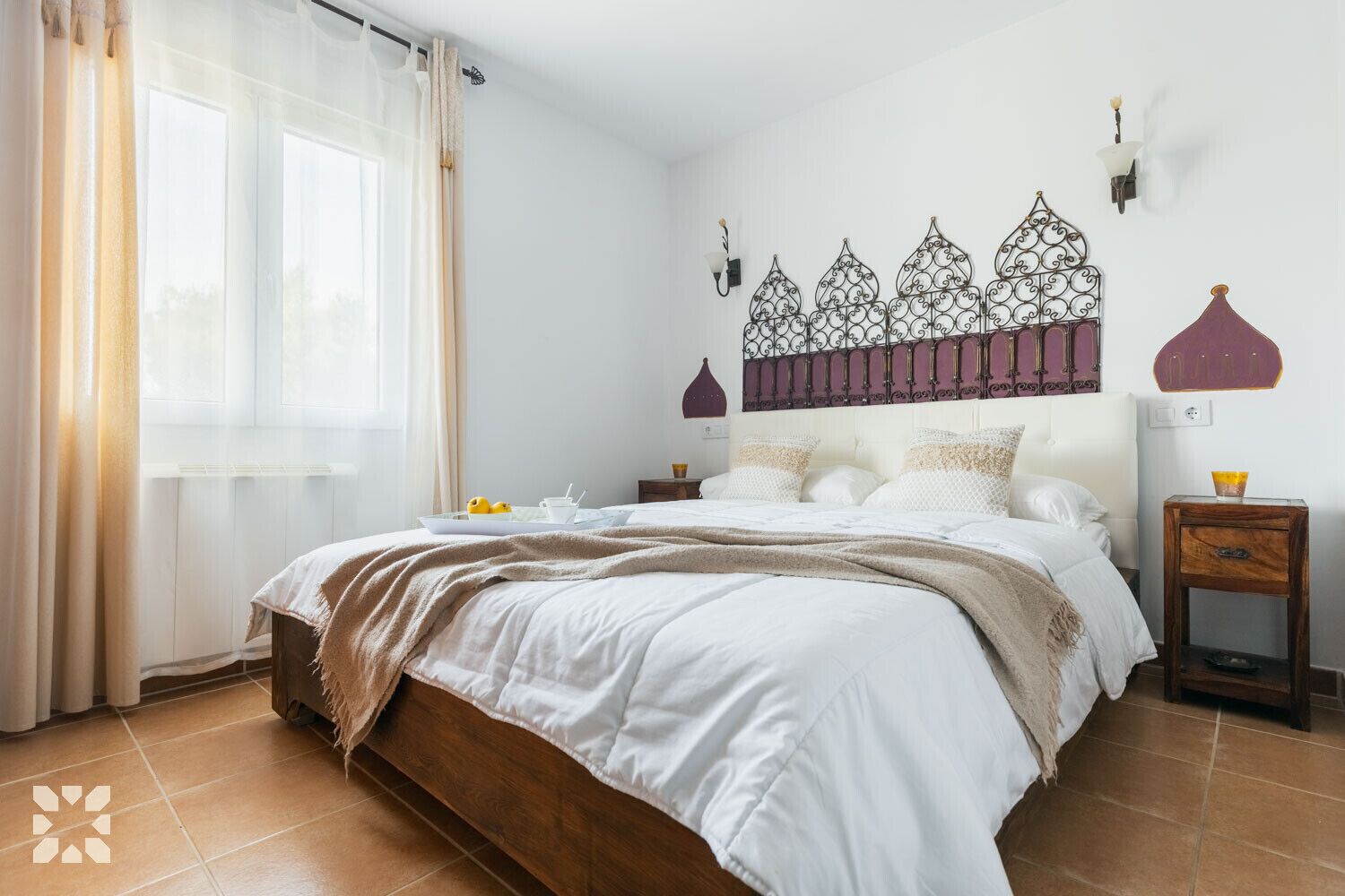 A bedroom with a bed, a nightstand, a wall-mounted lamp, and a window with curtains.