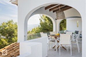A terrace with a table and chairs, a hanging light fixture, and a view of greenery.