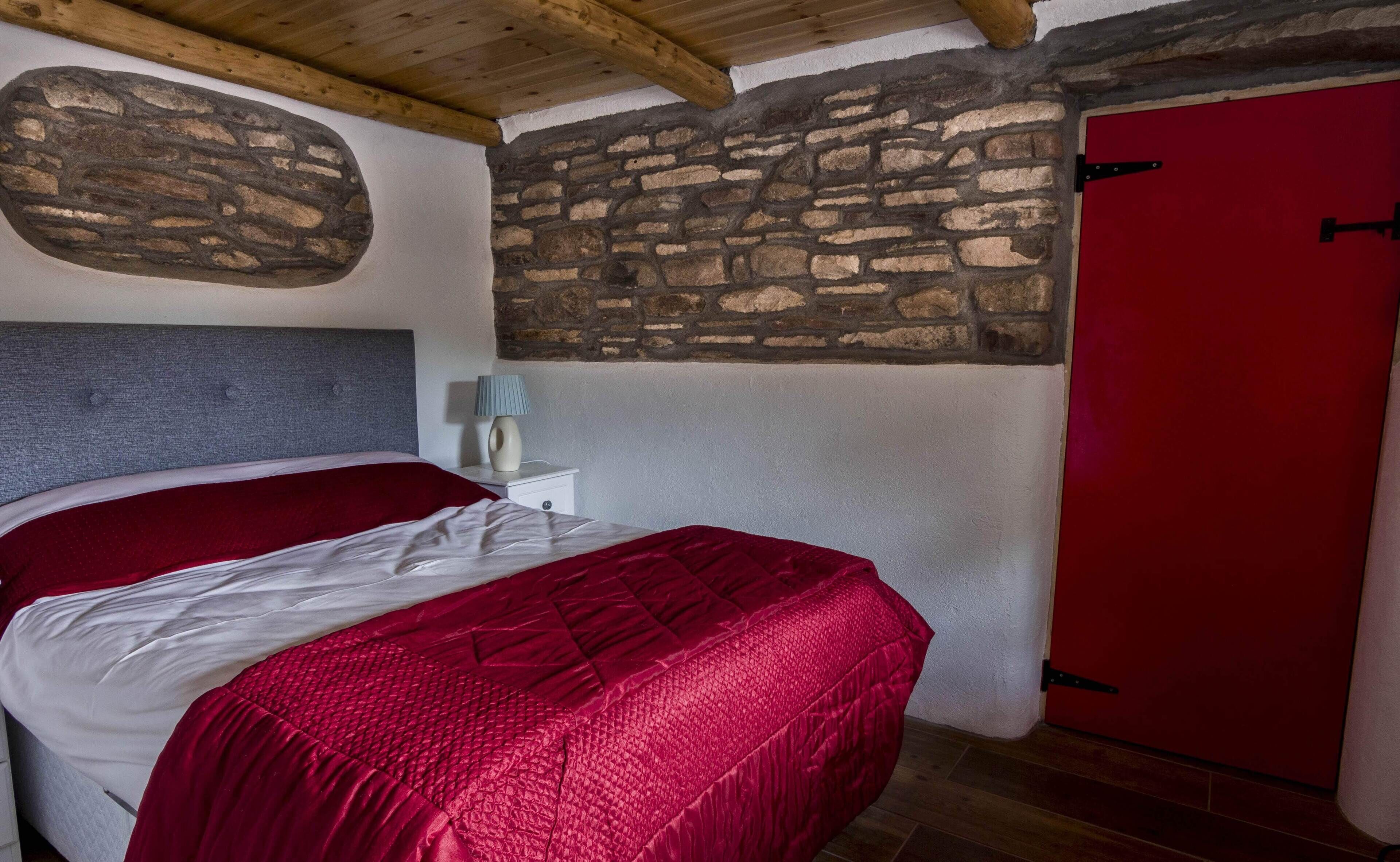 A bedroom with a stone wall, a red door, and a bed with red bedding.