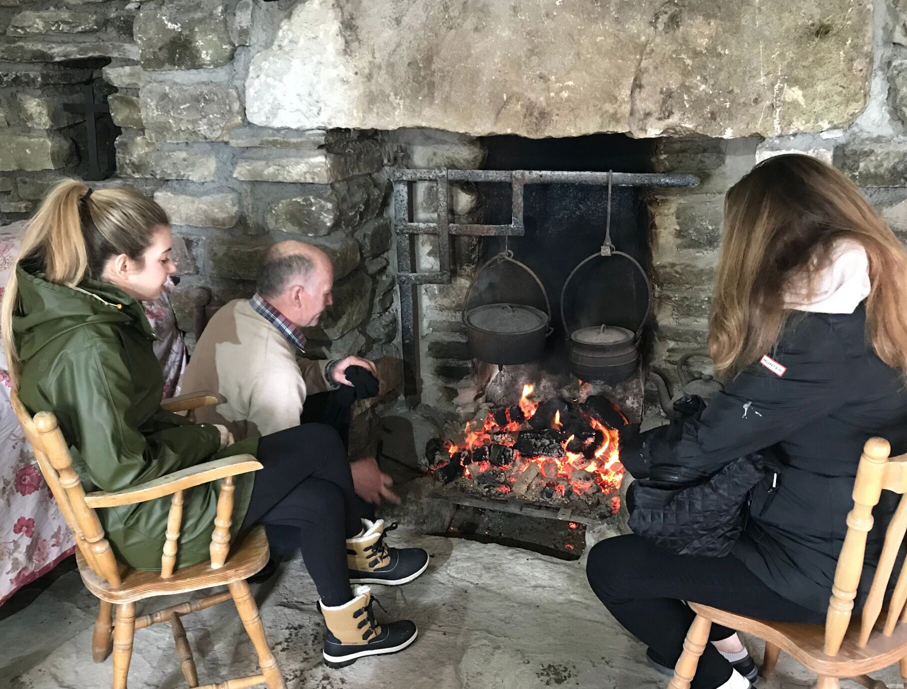 Three people seated around a stone fireplace with pots hanging above it.