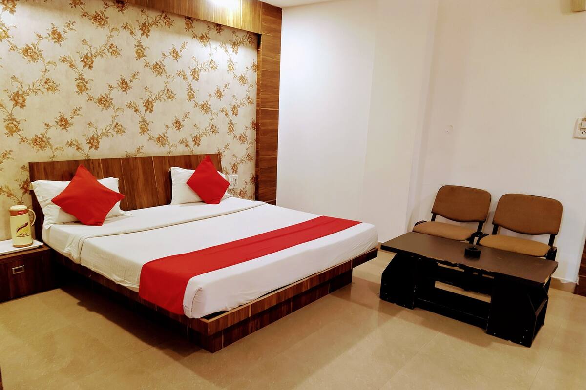 Ein Hotelzimmer mit einem hölzernen Bett, zwei roten Kissen, einer weißen Bettdecke, einer roten Decke, einem Nachttisch aus Holz, einer Kaffeemaschine und einem kleinen Tisch mit zwei Stühlen.