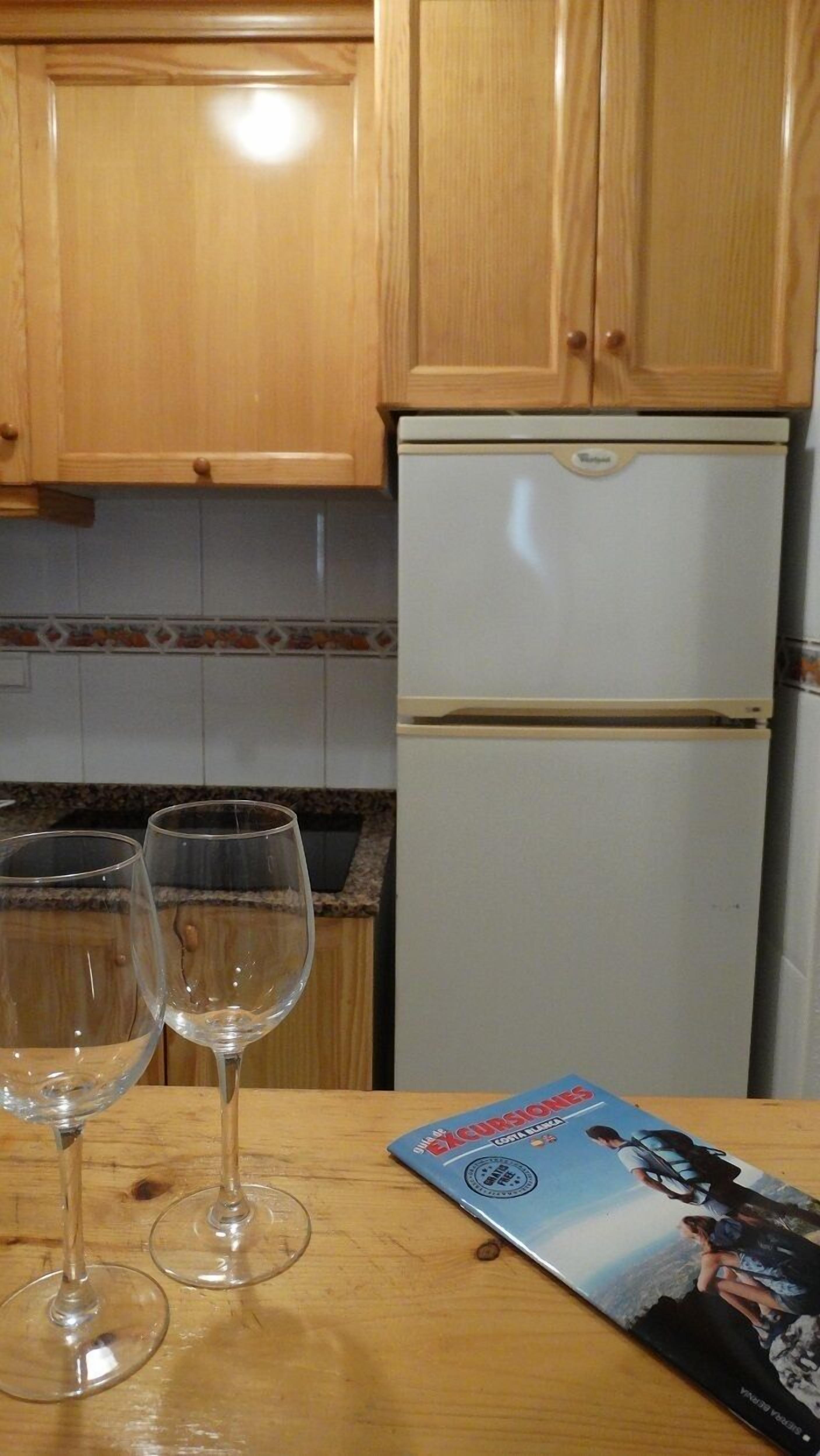 A kitchen with wooden cabinets and a white refrigerator. There are two empty wine glasses on the counter and a magazine with a travel theme.