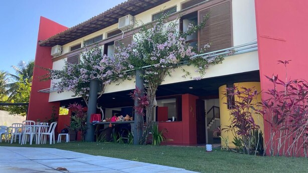 A two-story building with a red and beige facade, a covered patio area with outdoor seating, and a swimming pool in the foreground. - Beach House in Porto Seguro (Coroa Vermelha) 30 meters from the beach.  (BA)