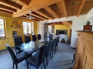 A dining area with a long table and chairs, wooden beams on the ceiling, and a TV in the background.