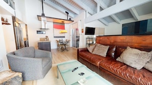 A spacious living area with a leather sofa, a patterned armchair, and a coffee table. There is a kitchen area with a refrigerator and a dining table.