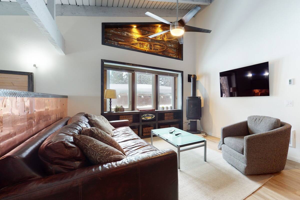 A living room with a brown leather sofa, a glass coffee table, a grey armchair, a large window, and a black wood-burning stove.