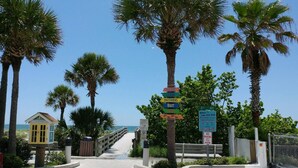 A beachside walkway with palm trees, a wooden bench, and a signpost with various directional signs.