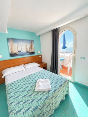 Une chambre d’hôtel avec un lit, un tableau représentant des voiliers, une vue sur la mer par la fenêtre et un parapluie bleu à l’extérieur.