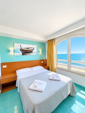A hotel room with a bed, a nightstand, a painting of a boat, and a view of the sea through large windows.