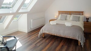 Ein Schlafzimmer mit schräg gedachter Decke, Dachfenstern, einem Bett mit vielen Kissen, einem Nachttisch mit Lampe und einem Sessel.