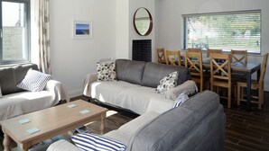 Ein Wohnzimmer mit einer Sofagarnitur, einem Couchtisch, einem Esstisch mit Stühlen, einem Kamin und einem großen Fenster mit Jalousien.