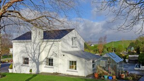 Une maison blanche à deux étages, avec une petite serre attenante, entourée d’une pelouse bien entretenue et d’un paysage vallonné en arrière-plan.