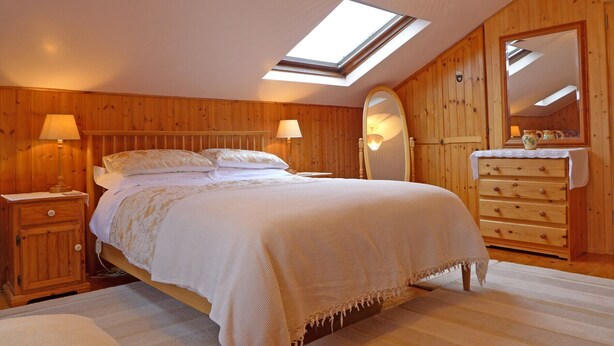 Une chambre avec une lucarne, des murs en bois, un lit avec du linge de lit blanc, deux tables de chevet avec des lampes et une commode avec un miroir.
