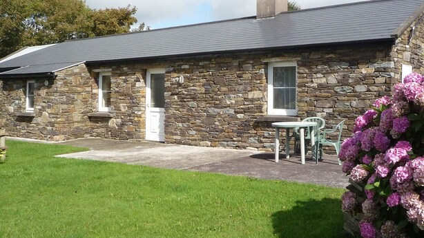 A stone building with a grey roof, a door, and a window. There is a green lawn and a bush with pink flowers. - Lakeside Lodge Bantry (Bantry)