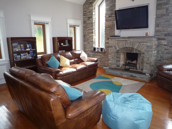 A living room with leather sofas, a stone fireplace, a flat-screen TV, and a bookshelf. - Seaview Holiday Home Bantry (Gortnakilly)