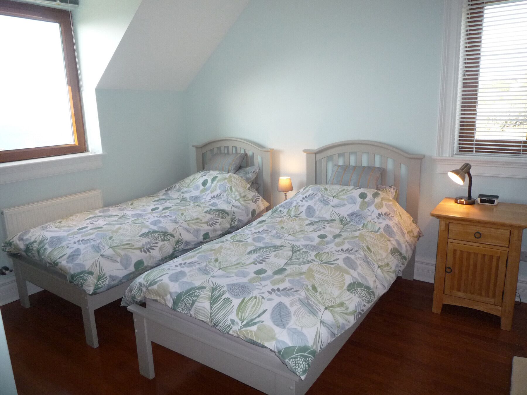 A bedroom with two single beds, a bedside table with a lamp, a radiator, and a window with blinds.