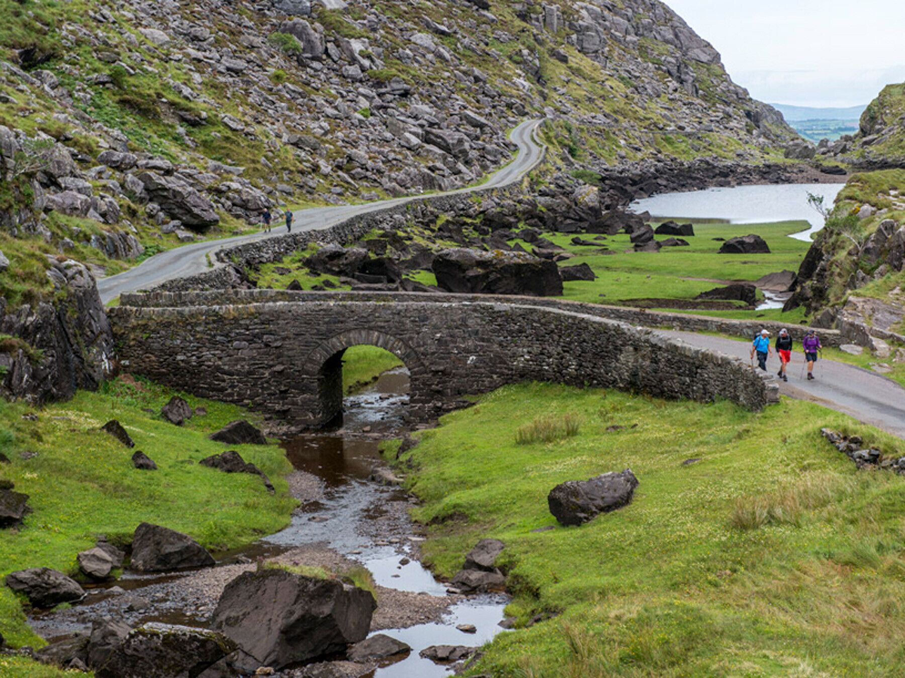 Un pont de pierre enjambe un ruisseau dans un paysage rural, avec des champs couverts d’herbe et un terrain rocheux.