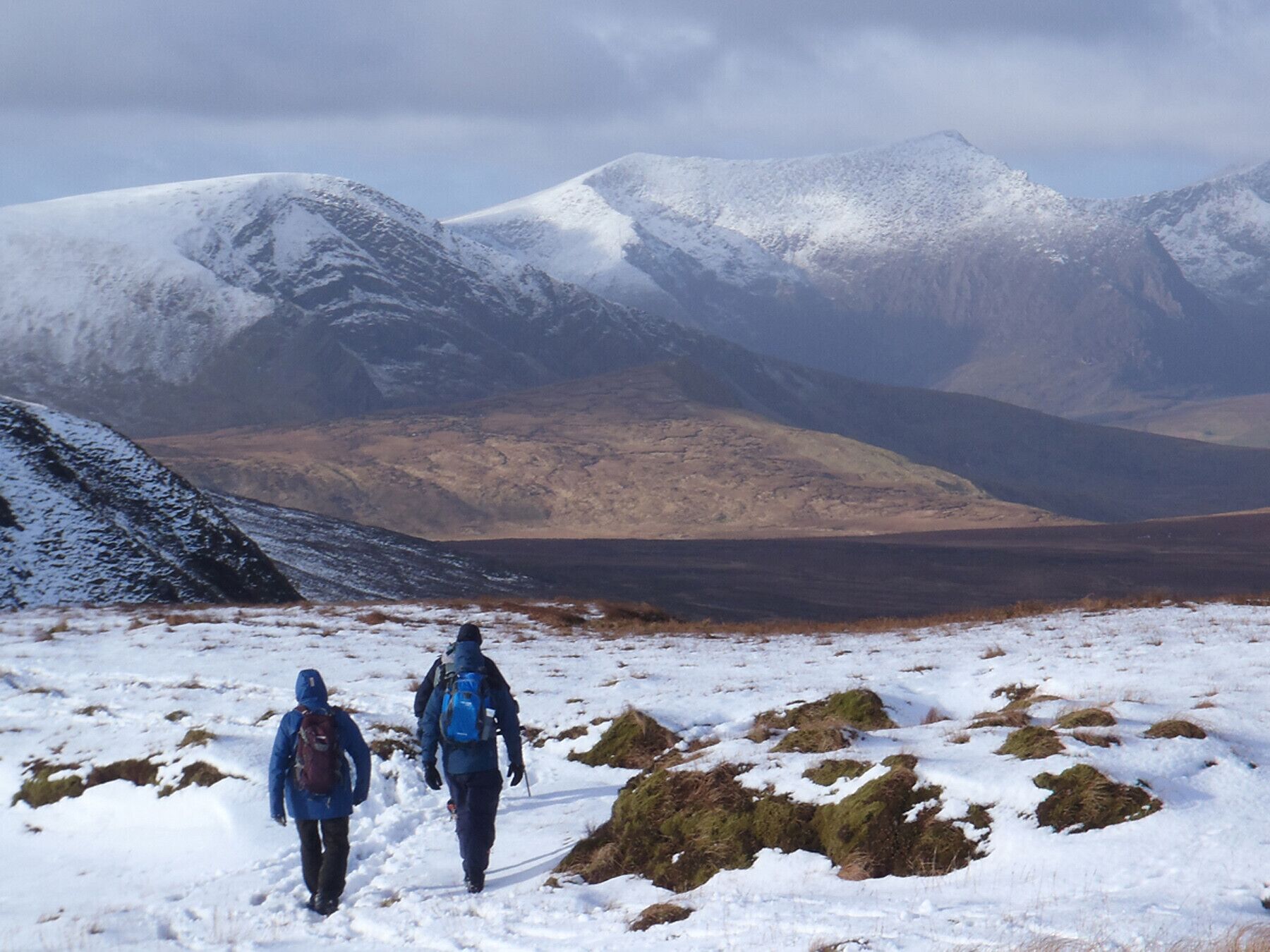 Two hikers on a snow-covered trail with mountains in the background.